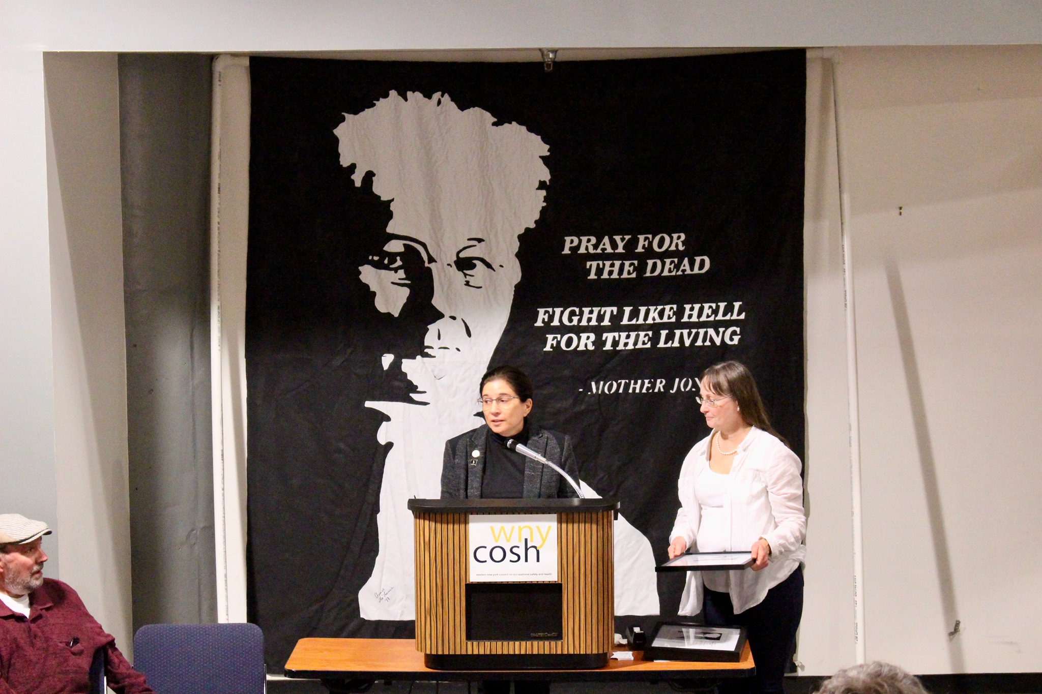 Two women speak in front of a Mother Jones banner