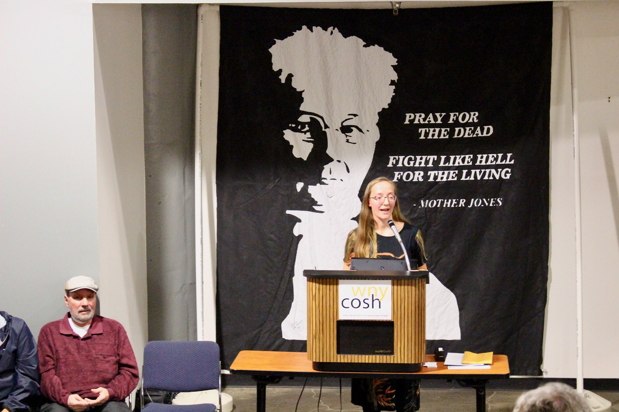 Picture of a woman speaking in front of a banner with Mother Jones on it