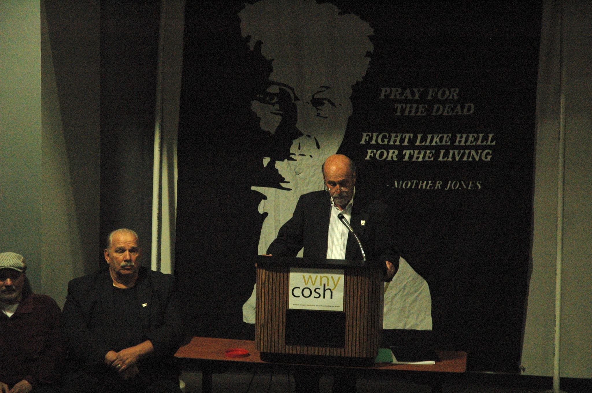 Picture of a man giving a speech in front of a WNYCOSH banner with Mother Jones on it